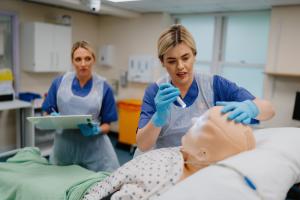 Two people in blue medical scrubs, gloves, and clear plastic aprons practise a clinical procedure on a realistic mannequin lying on a hospital bed, with one holding a syringe‑like instrument and the other observing with a clipboard in a clinical training room.