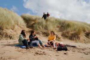 Three students sitting on the sand chatting and two students are walking towards them through the dunes behind them