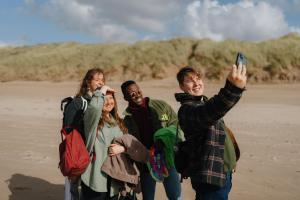 Pedwar myfyriwr ar lan y môr - mae un yn gafael mewn ffôn symudol ac yn cymryd llun o'r criw