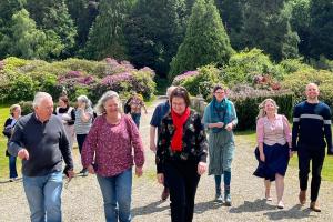 A group of people walking, all in conversation and smiling. Background of trees and flowering shrubs.