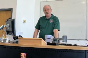 Vic wears a green ISWE polo shirt and stands behind a lectern 