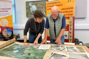 Dau ddyn yn sefyll dros fwrdd yn edrych ar rai dogfennau printiedig