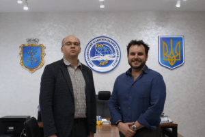 Two individuals standing in an office with three emblems on the wall behind them. The central emblem features a globe and text in Cyrillic, while the other two emblems display different symbols