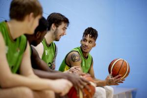 Myfyrwyr yn eistedd ar y meinciau yn sgwrsio yn un o'r neuaddau chwaraeon yng Nghanolfan Brailsford. Mae un yn gafael mewn pêl fasged.