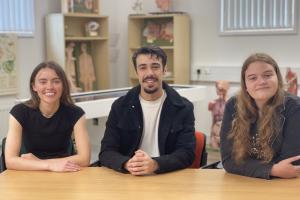 Three individuals seated at a wooden table in a classroom or lab setting, with anatomical models and educational charts visible in the background.