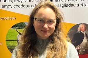 Isabel, who is wearing a grey jumper, is smiling and standing in front of an ISWE banner