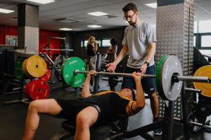 A person is weightlifting at the gym and another person is assisting