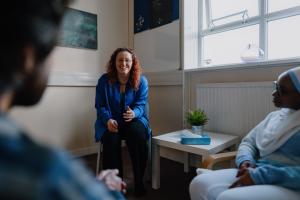 Three persons in a room, two are listening to the main speaker who is smiling 