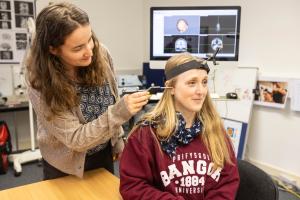 A Psychology lecturer using a neuronavigation pointer to localise a stimulation site on a student’s scalp during preparation for a transcranial magnetic stimulation (TMS) session, with brain images displayed on a computer screen in the background.