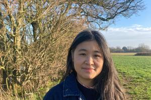 person â gwallt tywyll hir yn sefyll yn yr awyr agored wrth ymyl rhes o goed di-ddail ar ddiwrnod heulog. Maent yn gwisgo siaced denim dywyll. Y tu ôl iddynt ceir cae gwyrdd agored yn ymestyn i’r pellter o dan awyr las gyda chymylau gwasgaredig