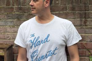 A person wearing a white T‑shirt with blue cursive text reading ‘The Hard Quartet’ stands in an outdoor garden area. They are positioned in front of a brick wall with green foliage and trees visible above and around it. A wooden shelving unit holding dishes is to the left. The person faces slightly to the side, with their arms relaxed by their sides.
