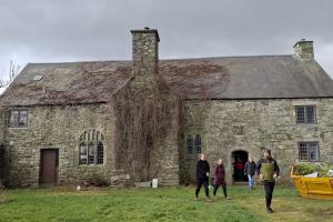 Ffotograff o dŷ carreg hir gyda simnai fawr yng nghanol y ffasâd. Yn y blaendir, mae sgip melyn a sawl person yn cerdded i ffwrdd o'r tŷ.