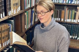 Woman in grey jumper and glasses standing in a library, reading a book. 