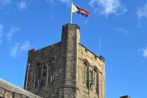 “Stone tower of a historic building with a pride flag and university emblem flying at the top against a bright blue sky