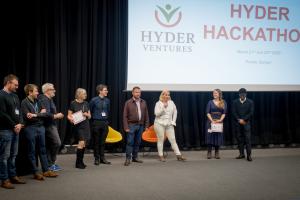 A group of people stand in a row on a stage during the Hyder Ventures Hackathon at Pontio, Bangor. Two participants hold certificates, and another stands near a microphone while addressing the audience. A large screen behind them displays the event name and dates.