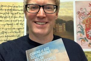 Mae Shaun yn sefyll, yn gwenu, o flaen baner ISWE, yn dal ei lyfr.