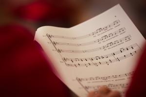 Close-up photograph of printed sheet music held at an angle, showing musical notation on multiple staves. The page is partially framed by out-of-focus red fabric in the foreground, with a softly blurred background