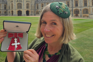 Person standing on a lawn in front of a historic stone building, holding an open presentation case displaying a silver medal with a red ribbon.