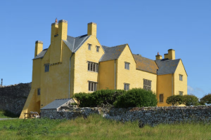 Photograph of a Tudor house with lots of gables and chimneys, with the walls painted bright yellow, set against a blue sky.