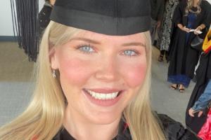 Woman with blond hair in black graduation outfit, with flat cap