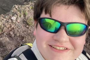 a picture of a student (aiden) looking into the camera and taking a picture. the student is wearing reflective sunglasses, and is smiling. the student has a rocky terrain behind him