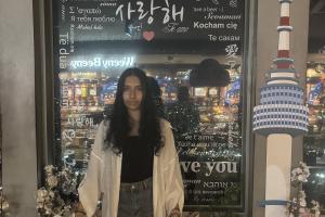 an image of a student (aneesha) standing infront of a building. the student is looking at the camera. behind her are some flowers and a window. she is wearing a white shirt and grey trousers