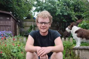 an image of a student (jem) sitting down to take a photo. the student is smiling, and he has his hands crossed over each other casually. the student is in a garden background, as has foliage around him. there is also a dog in the background