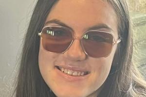 an image of a student (kayleigh) looking at the camera and smiling. the student is wearing sunglasses, and she is in a neutral room with a window behind her