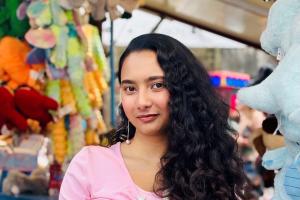 a picture of a student (rafia) in a pink shirt looking at the camera. the student is in a fair and is surrounded by fairground prizes in the background. the student is putting one hand on her hip and she is smiling