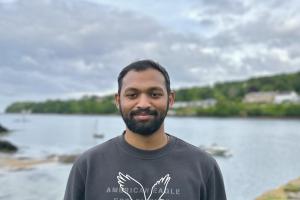 an image of a student (steffan) wearing a grey jumper with an eagle on it. the student is looking at the camera and smiling. the student is outside and has a scene of nature (river and grass) behind him
