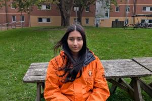 An image of Aneesha from campus life wearing an orange campus life coat. She is sitting on a bench outside 