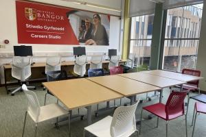 Stiwdio Gyrfaoedd ym Mhrifysgol Bangor, gyda chyfrifiaduron ar hyd un wal a byrddau a chadeiriau lliwgar wedi'u trefnu ar gyfer gwaith grŵp.