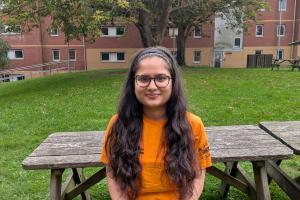 An image of Ishneha from campus life wearing an orange campus life tshirt. She is sitting on a bench outside 