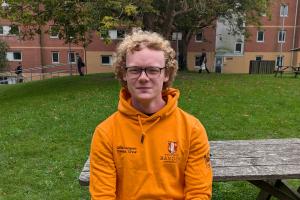 An image of Jem from campus life wearing an orange campus life hoodie . He is sitting on a bench outside 