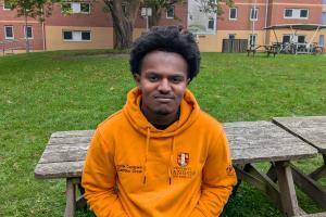 An image of Josh from campus life wearing an orange campus life hoodie. He is sitting on a bench outside 