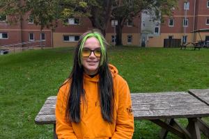 An image of Kayleigh from campus life wearing an orange campus life hoodie. She is sitting on a bench outside 