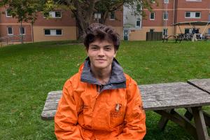 An image of Luke from campus life wearing an orange campus life coat. He is sitting on a bench outside 