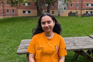An image of Rafia from campus life wearing an orange campus life tshirt. She is sitting on a bench outside 