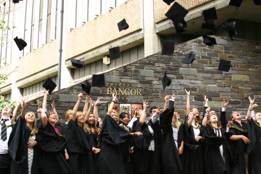 Students celebrating by throwing their caps in the air after their graduation ceremony with the Bangor University sign in the background