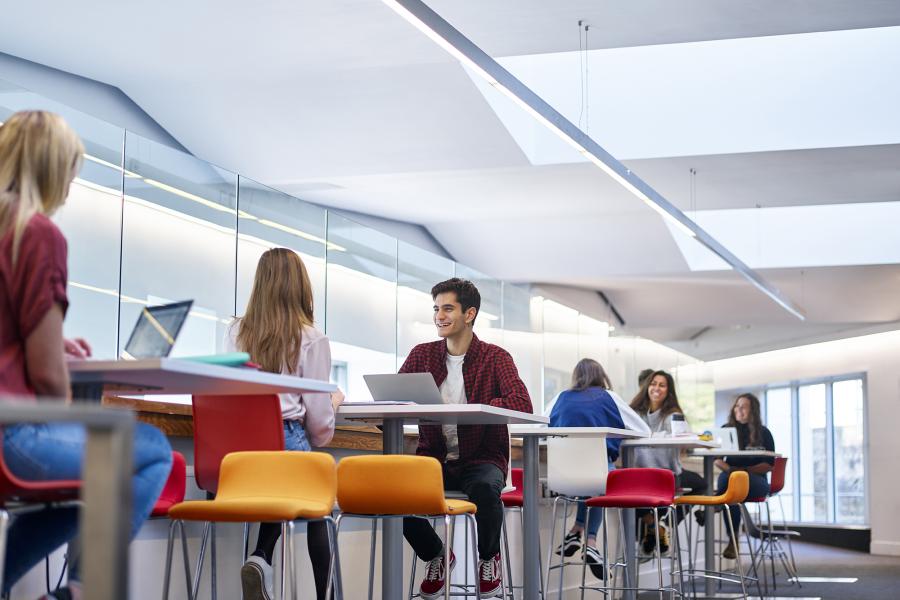 students in social learning space chatting, laptops and books on tables