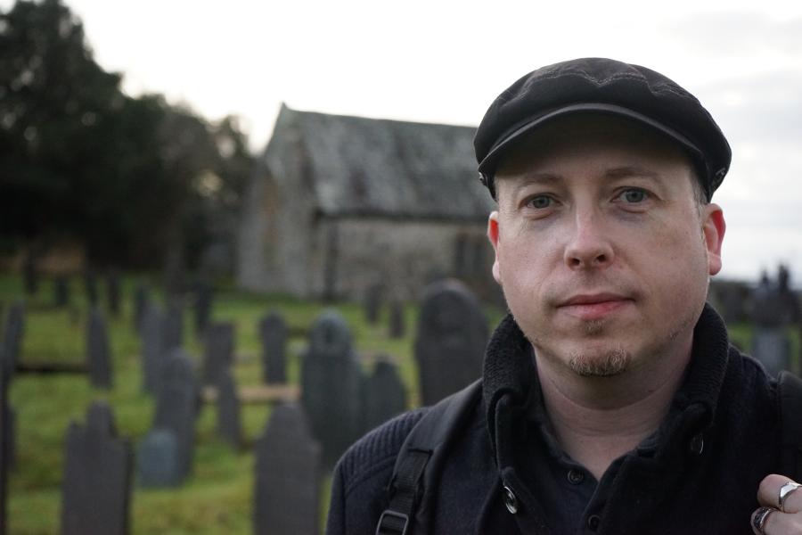 Man in flat cap stands, in the distance is a small church.