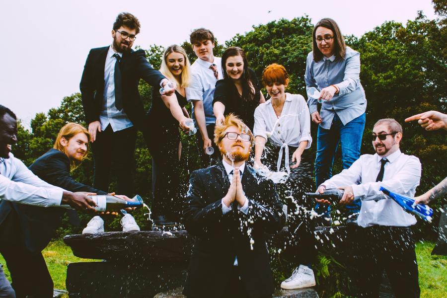 A crazy assemblage of young people pointing as one is showered with champagne