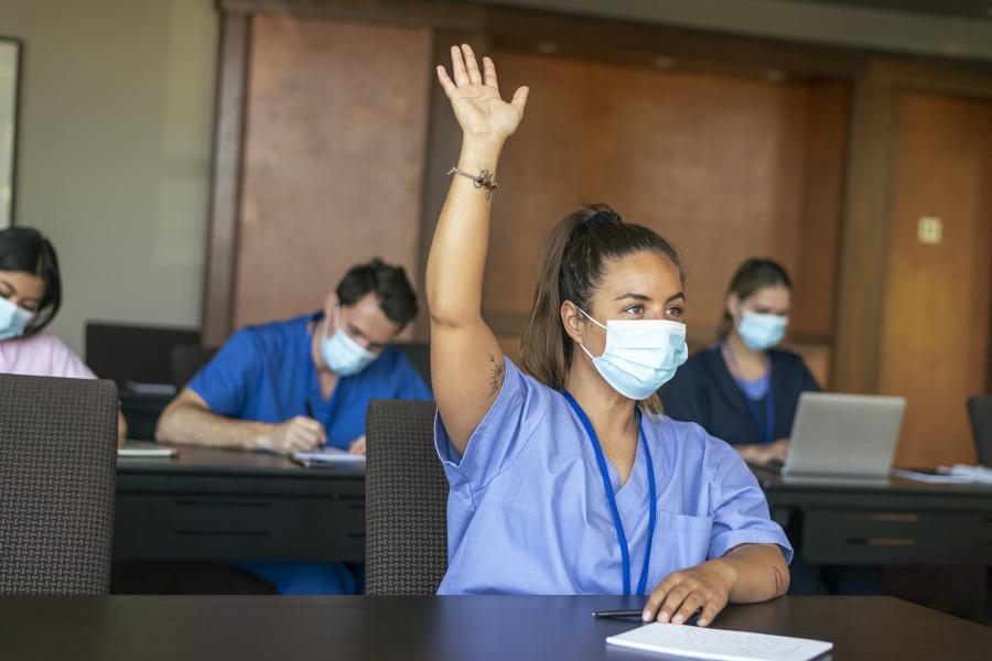 A woman in 'scrubs' and mask holds up her hand in a classroom, others are writing