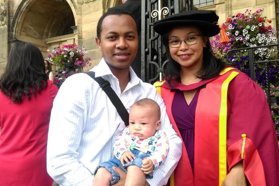 Family group of woman in  red graduation gown and husband and baby.