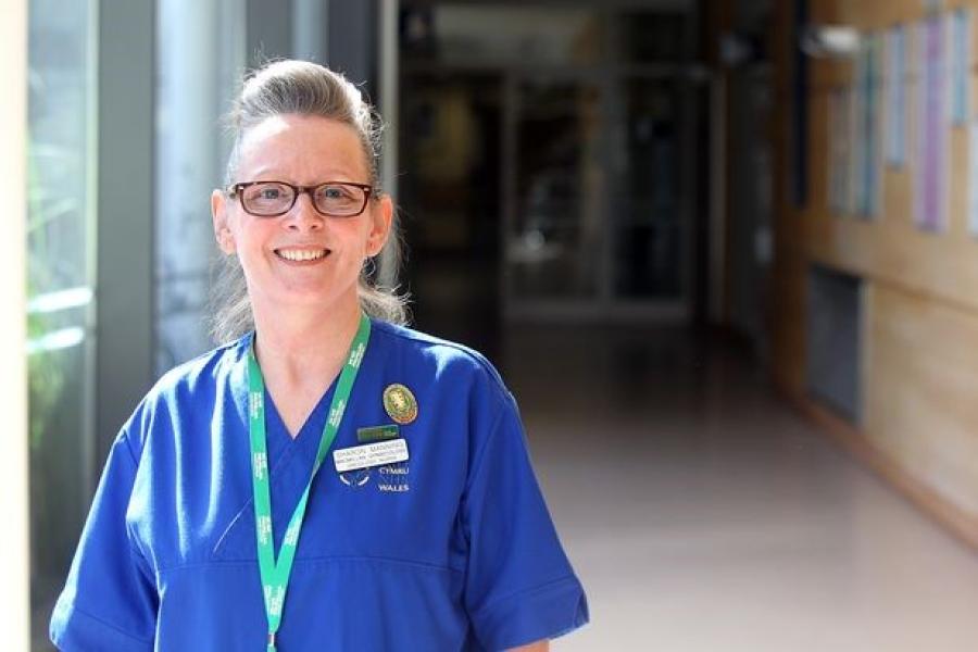 Woman in scrubs ans specatcles smiles into camera from left of frame