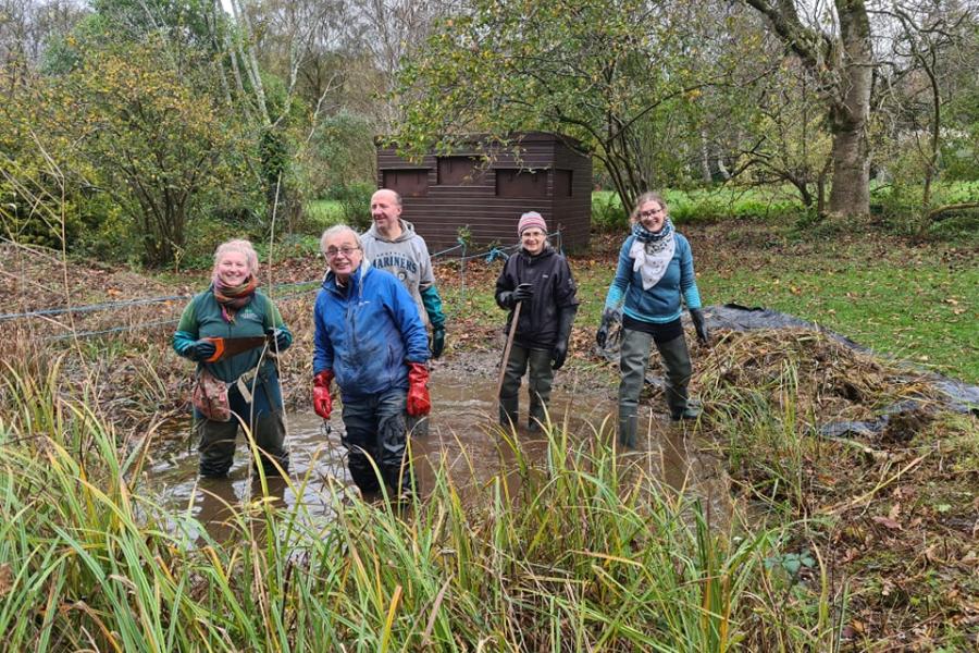 A group of five staff and volunteers stand in a pond, wearing waders and looking towards the camera, at Treborth Botanic Garden