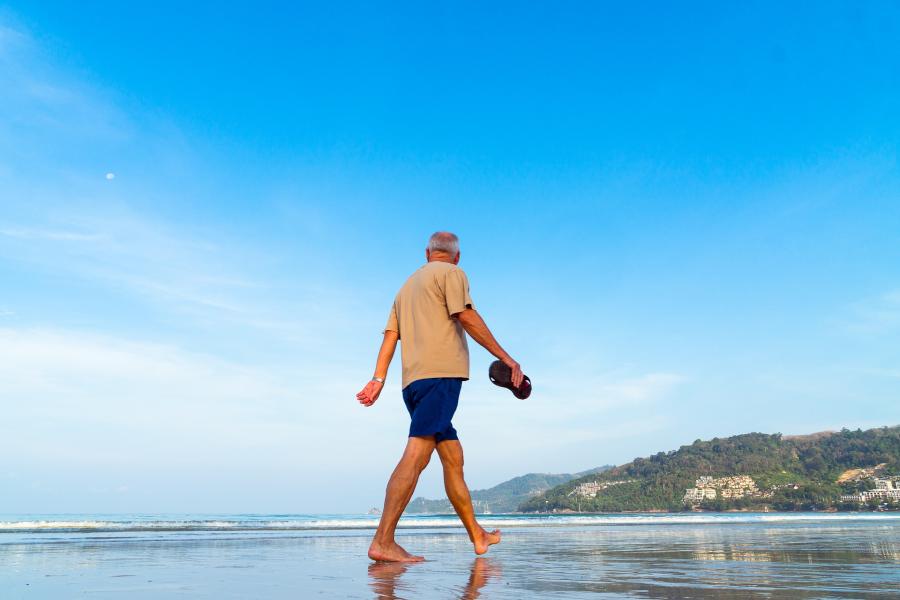 Man in shorts and T shirt walks on beach, carrying shoes