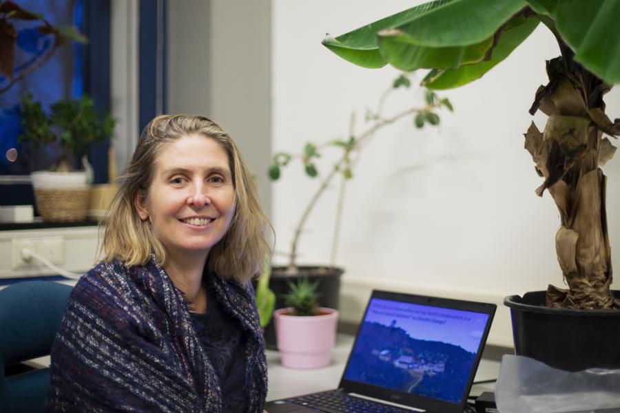Professot Julia Jones, with blonde shoulder length hair smiles into camera, she is seated in front of a laptop computer and large tropical looking houseplant.