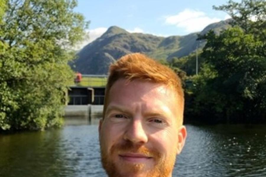 Red haired and bearded, head and shoulderds of dan Curran stands in front of water, with mountain in distance.