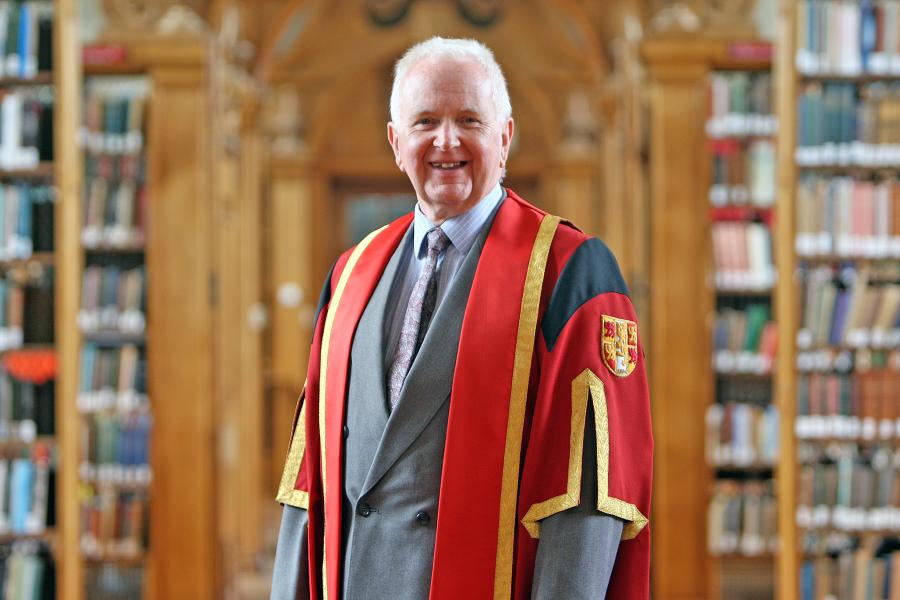 White haired man stands in red graduation gown in Bangor University's Shankland library